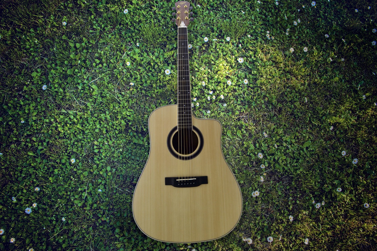 Acoustic guitar lying flat on lush green grass scattered with small white daisies, shot from directly above.