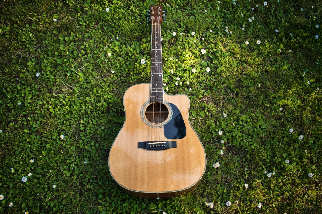 Acoustic guitar lying flat on lush green grass scattered with small white daisies, shot from directly above.