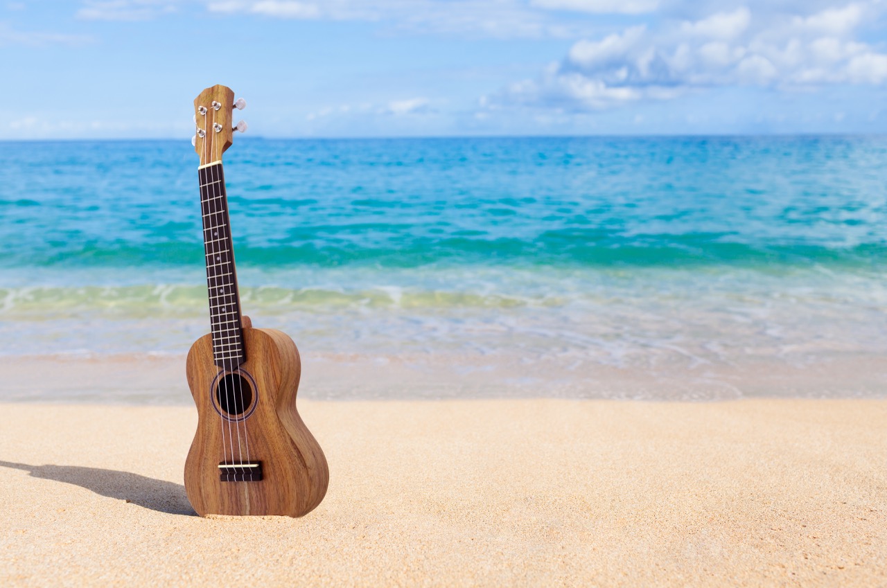 Wooden ukulele standing upright in the sand on a tropical beach with turquoise ocean waves and blue sky in the background.