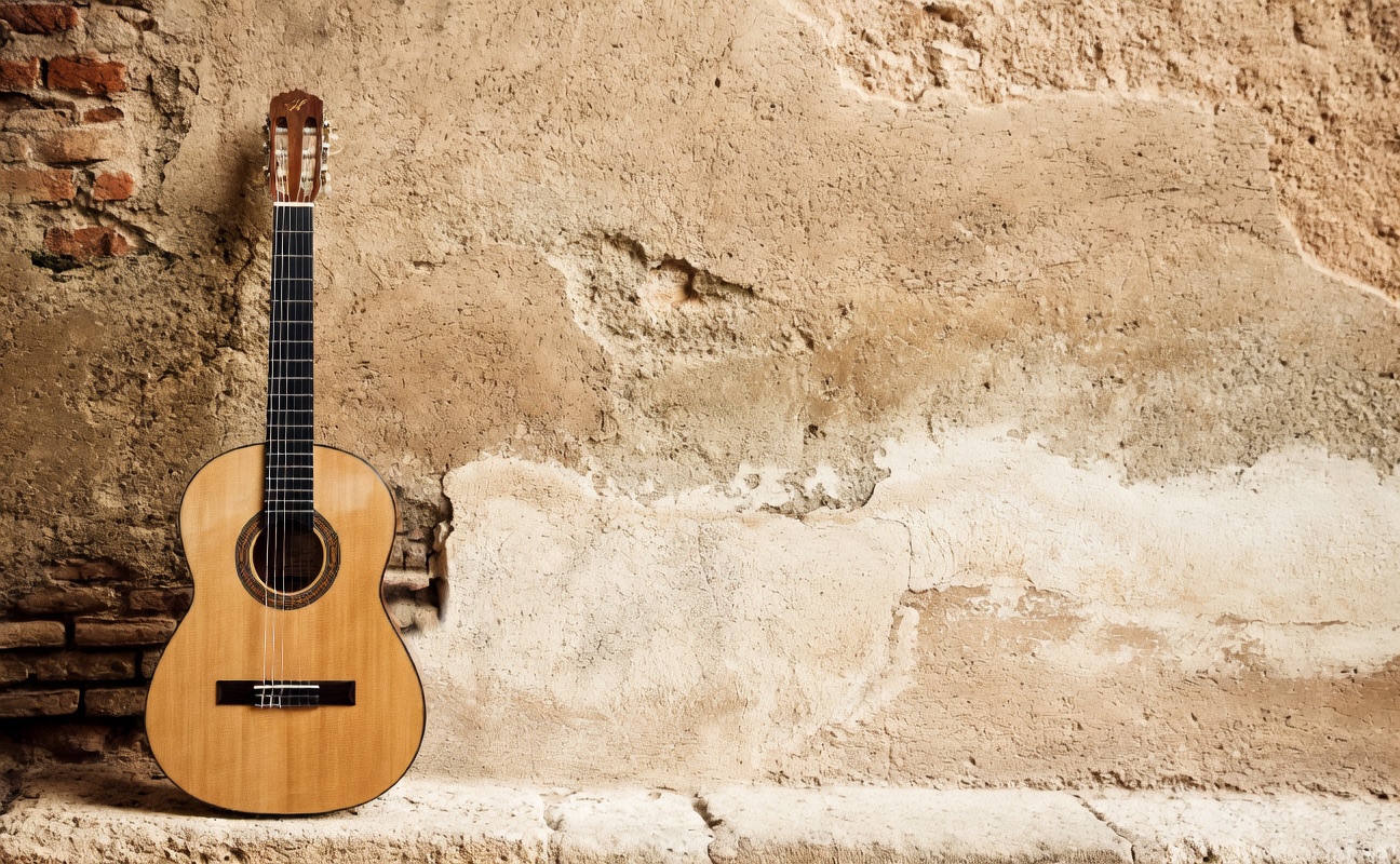Light-colored acoustic guitar leaning against an aged, peeling plaster wall with brick details at the base.