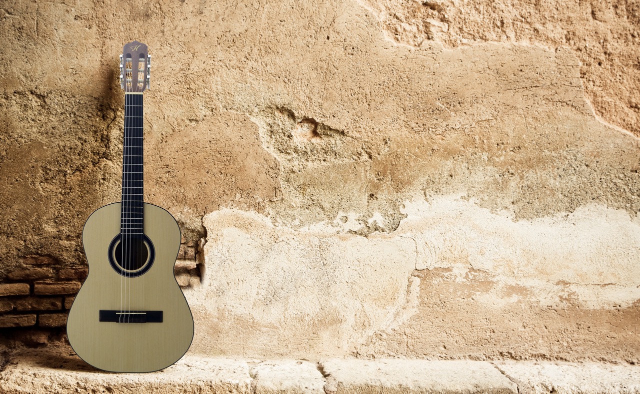 Light-colored acoustic guitar leaning against an aged, peeling plaster wall with brick details at the base.
