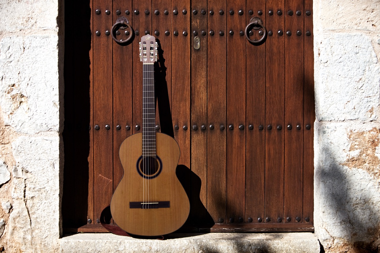 Classical acoustic guitar leaning against a large dark wooden studded door framed by white stone walls.