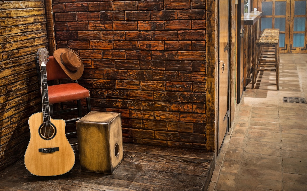 Acoustic guitar, wooden cajón percussion box, and brown cowboy hat on a leather chair in a rustic wooden bar interior.